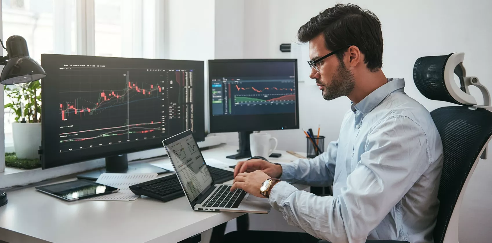 Workplace of trader. Young bearded trader wearing eyeglasses using his laptop while sitting in office in front of computer screens with trading charts and financial data