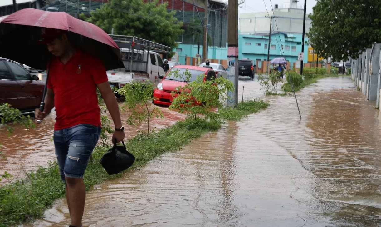 Hombre camin  con sombrilla y lonchera en mano,   en acera inundada en la    Capital.